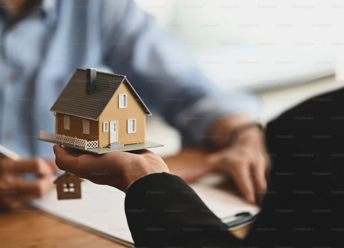 Khách hàng cầm mô hình căn nhà trao đổi với nhân viên văn phòng đang cầm bút ghi chú – minh họa cho dịch vụ mua bán nhà đất trọn gói an toàn và chuyên nghiệp